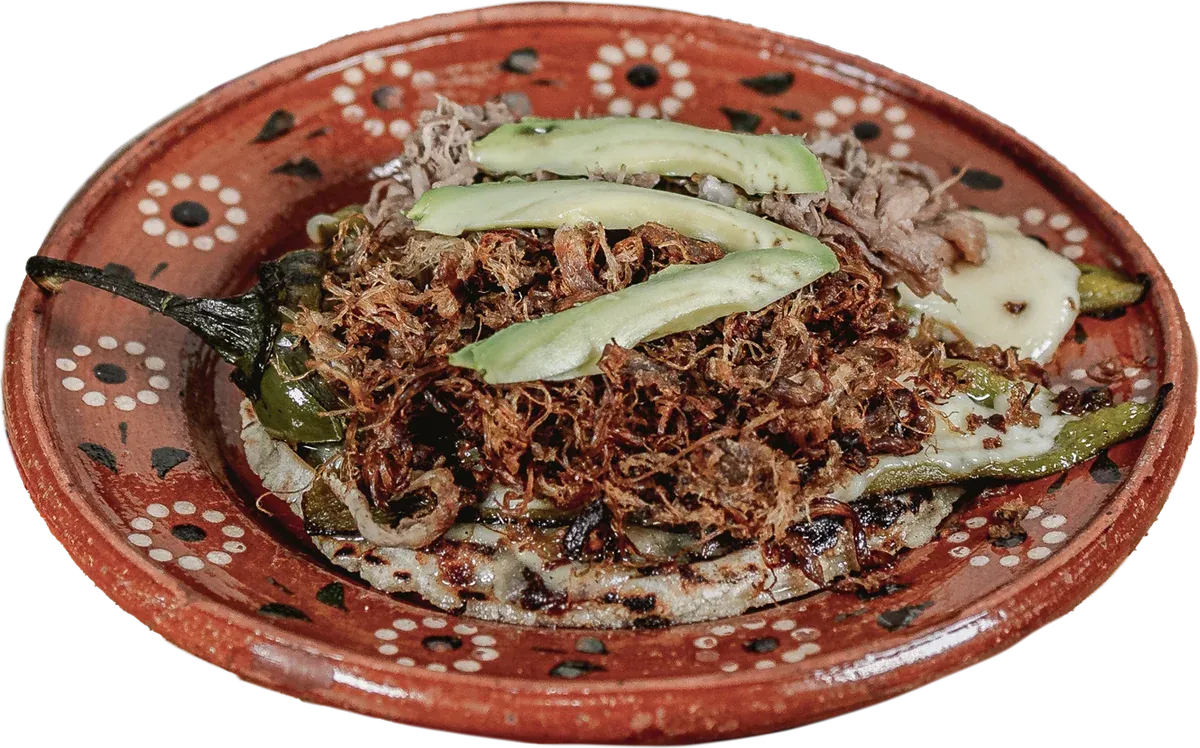 Pellizcada mexicana: tortilla de maíz con carne de borrego deshebrada, chile Anaheim asado y láminas de aguacate en plato de barro